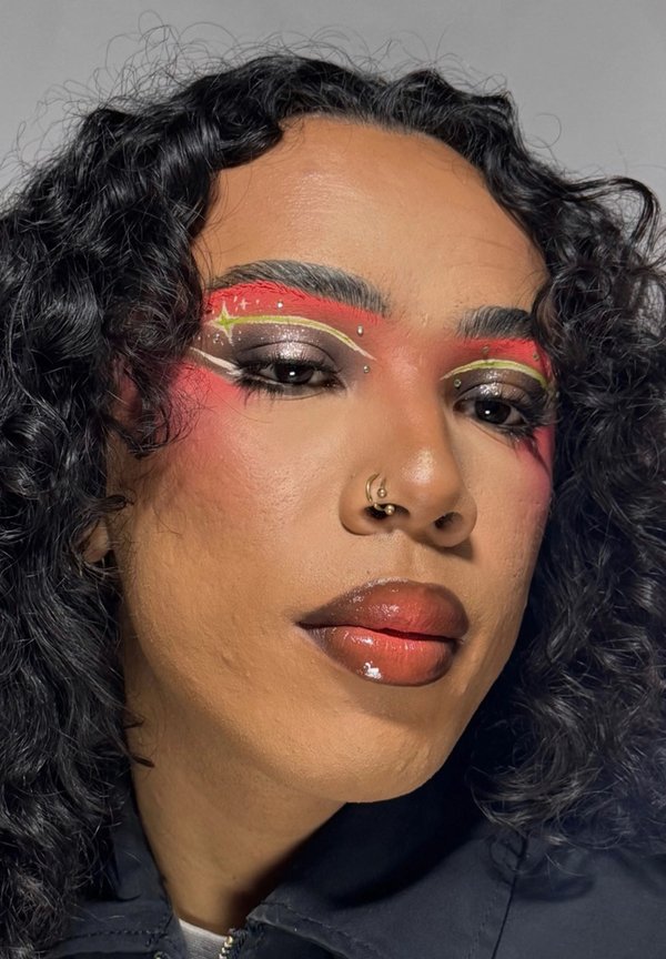 Person mit lockigem schwarzen Haar, trägt farbenfrohes Augen-Make-up mit roten, weißen und gelben Akzenten und glänzendem dunkelrotem Lippenstift, mit Nasenpiercings.