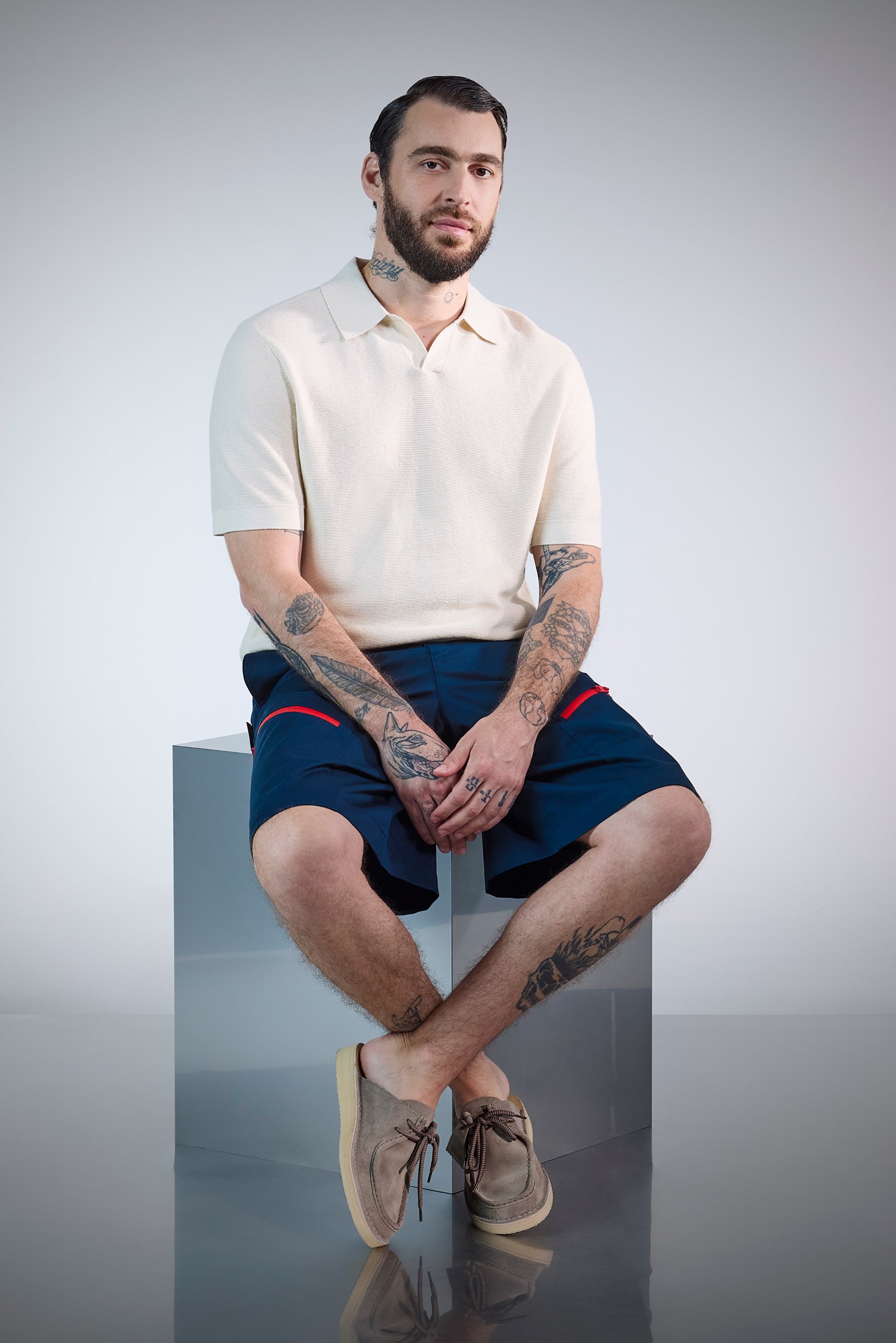 A man with tattoos sits on a silver pedestal, wearing a light polo shirt and navy shorts, complemented by brown shoes. Neutral background.