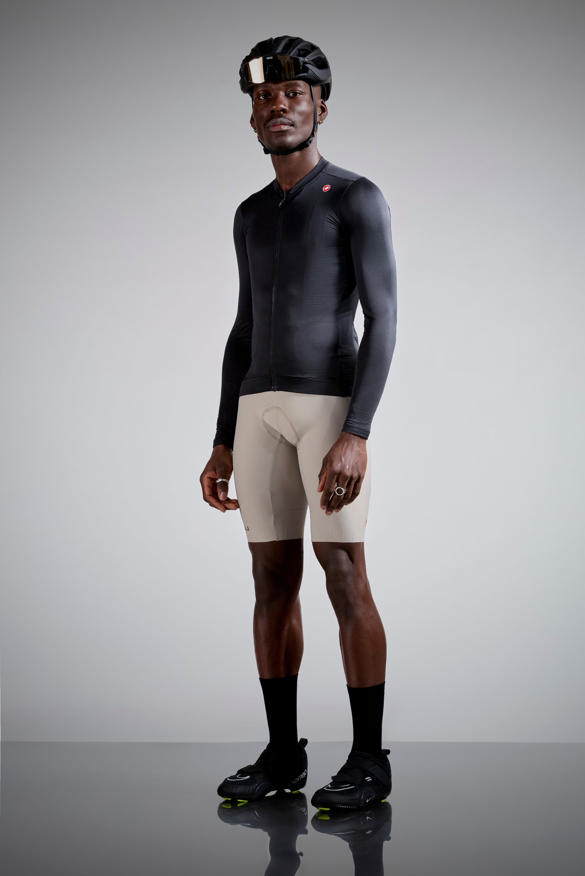 A cyclist stands confidently in a black long-sleeve jersey and beige shorts, wearing a black helmet and sleek cycling shoes.