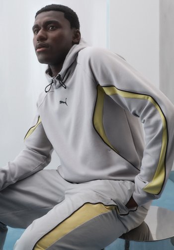 Млад мъж, облечен в светлосив екип на Puma с жълти райета, седнал и гледащ настрани на неутрален фон.