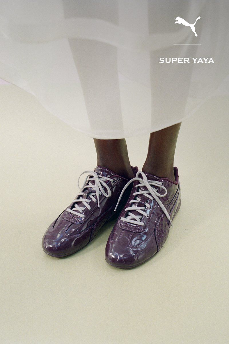 Person, die glänzende lila Puma Super Yaya Sneaker mit weißen Schnürsenkeln trägt, steht auf einem hellen Boden unter einem weißen fließenden Rock.