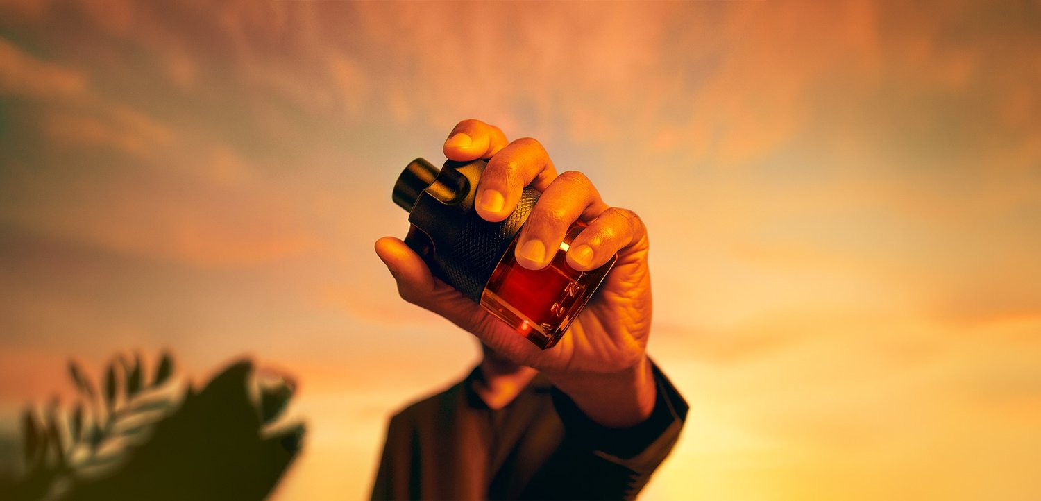 Hand hält eine dunkle Glasflasche mit schwarzem Verschluss vor einem warmen, orangefarbenen Sonnenuntergangshimmel.