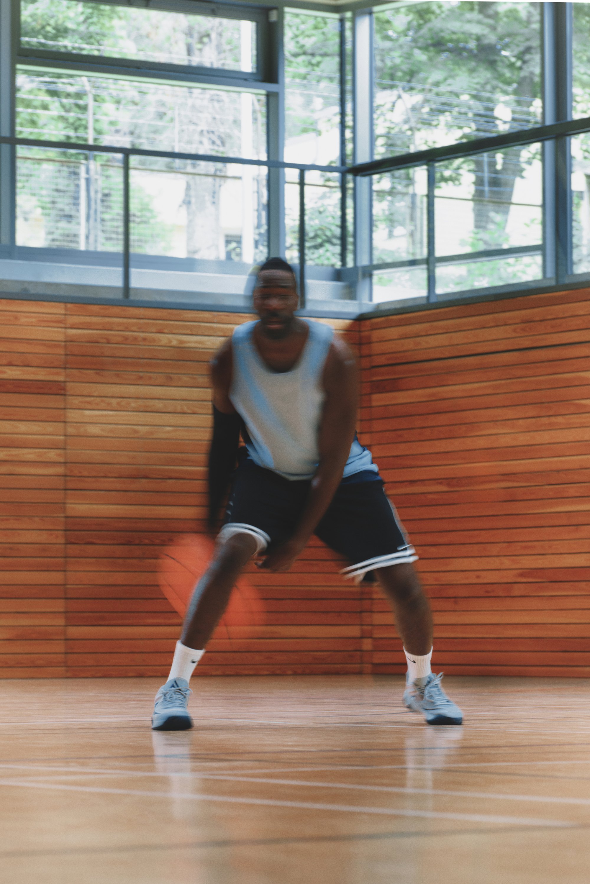 Basketballspeler dribbelend op een houten vloer, gekleed in een lichtblauw tanktop en zwarte shorts, met grijze sneakers, in een sportschoolsetting.