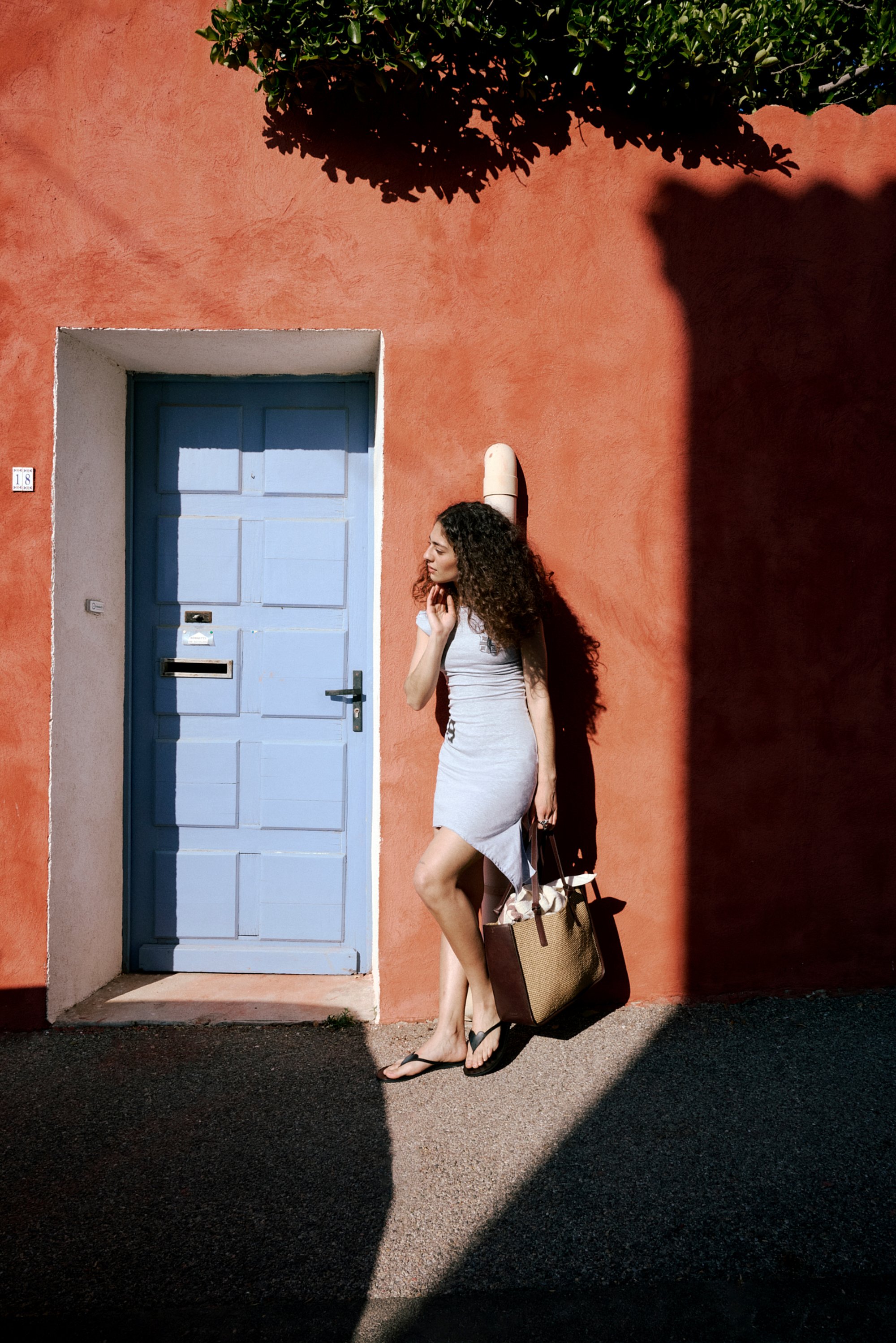 Una mujer con cabello rizado está de pie junto a una pared roja, al lado de una puerta azul claro, sosteniendo una bolsa grande mientras es iluminada por la luz del sol y sombras.