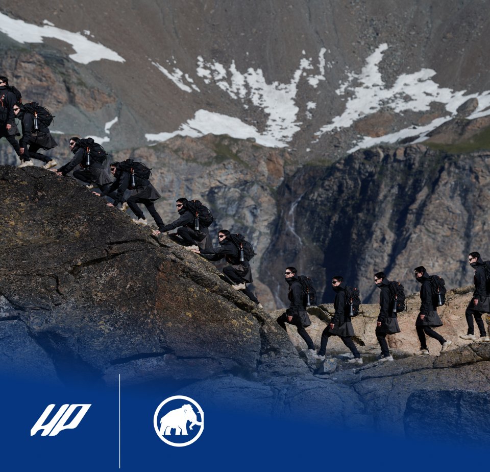 Mehrere Wanderer in schwarzen Jacken und Rucksäcken wandern einen felsigen Bergweg entlang, mit rauem Terrain und schneebedeckten Gipfeln im Hintergrund.