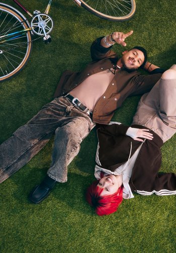 Dos jóvenes tumbados en la hierba junto a una bicicleta, uno con el pelo rojo sonriendo, el otro señalando hacia arriba y sonriendo.