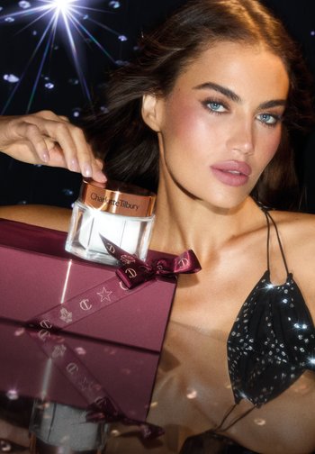 Clear jar of Charlotte Tilbury skincare product with a rose gold lid, positioned beside a burgundy gift box tied with a matching ribbon.