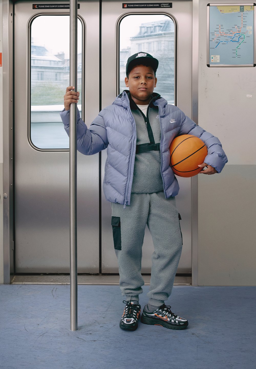 En pojke står i en tunnelbana med en ljuslila vadderad jacka, grå fleece-träningsdräkt med svarta detaljer och mönstrade sneakers, och håller en basketboll.
