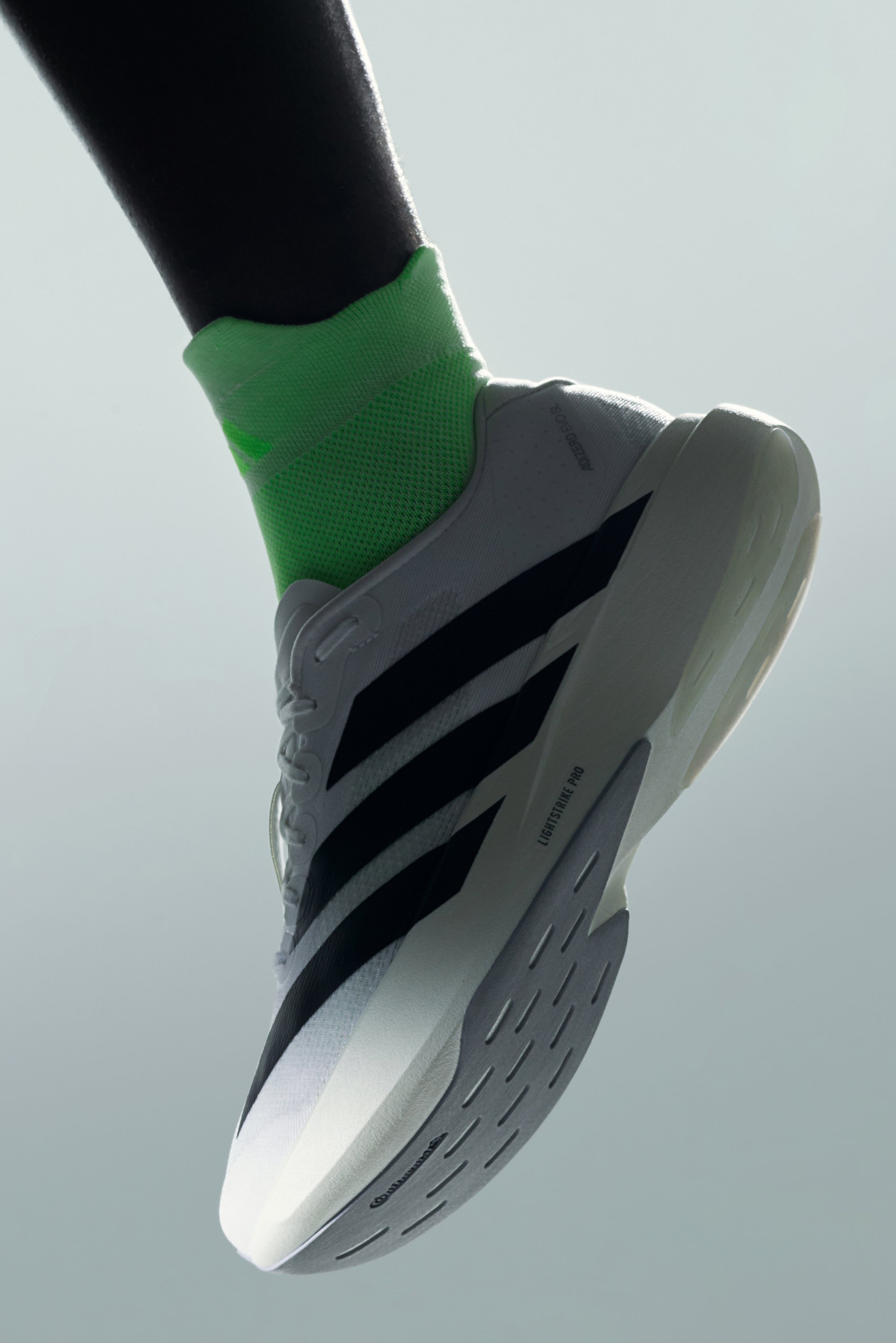Un primer plano de una zapatilla deportiva blanca con rayas negras, combinada con un calcetín verde brillante, sobre un fondo gris suave.