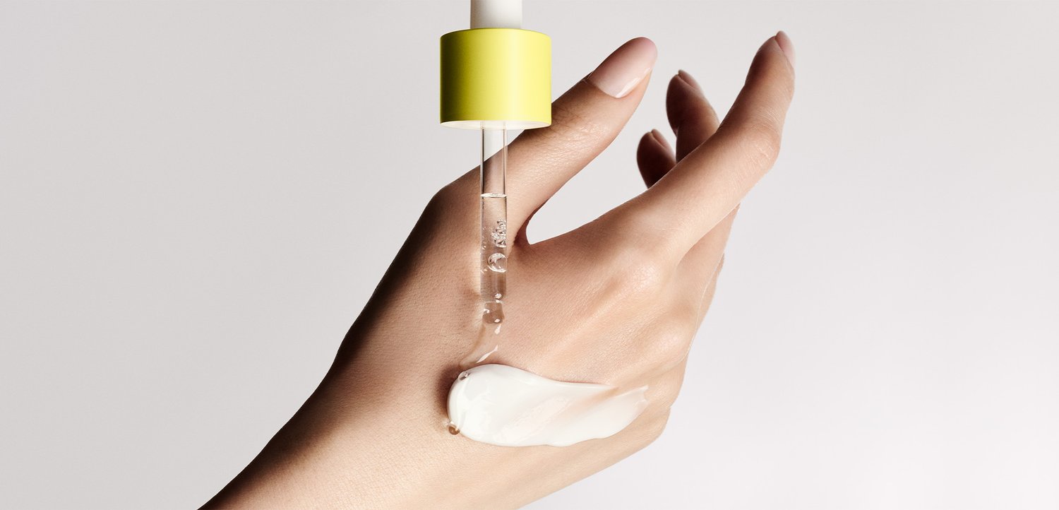 Hand with light skin tone holds fingers apart as a dropper with a yellow top dispenses liquid onto lotion spread on the back of the hand.