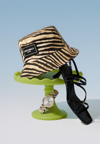 Tiger-striped bucket hat with "Just Cavalli" label, black strappy high heel, and two-tone metal wristwatch on a green cake stand.