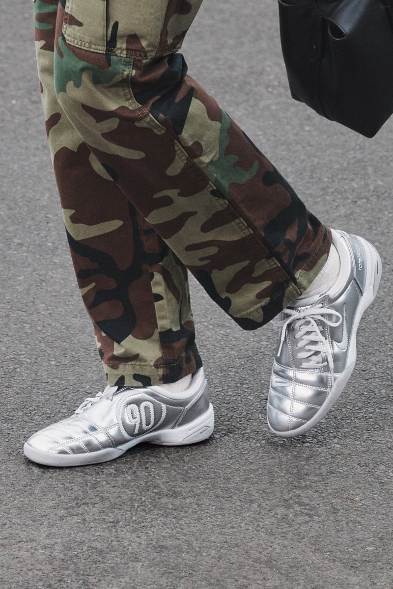 Des sneakers argentés avec une surface texturée et un '90' brodé sur le côté, associés à un pantalon camouflage, exposés sur un pavé gris.