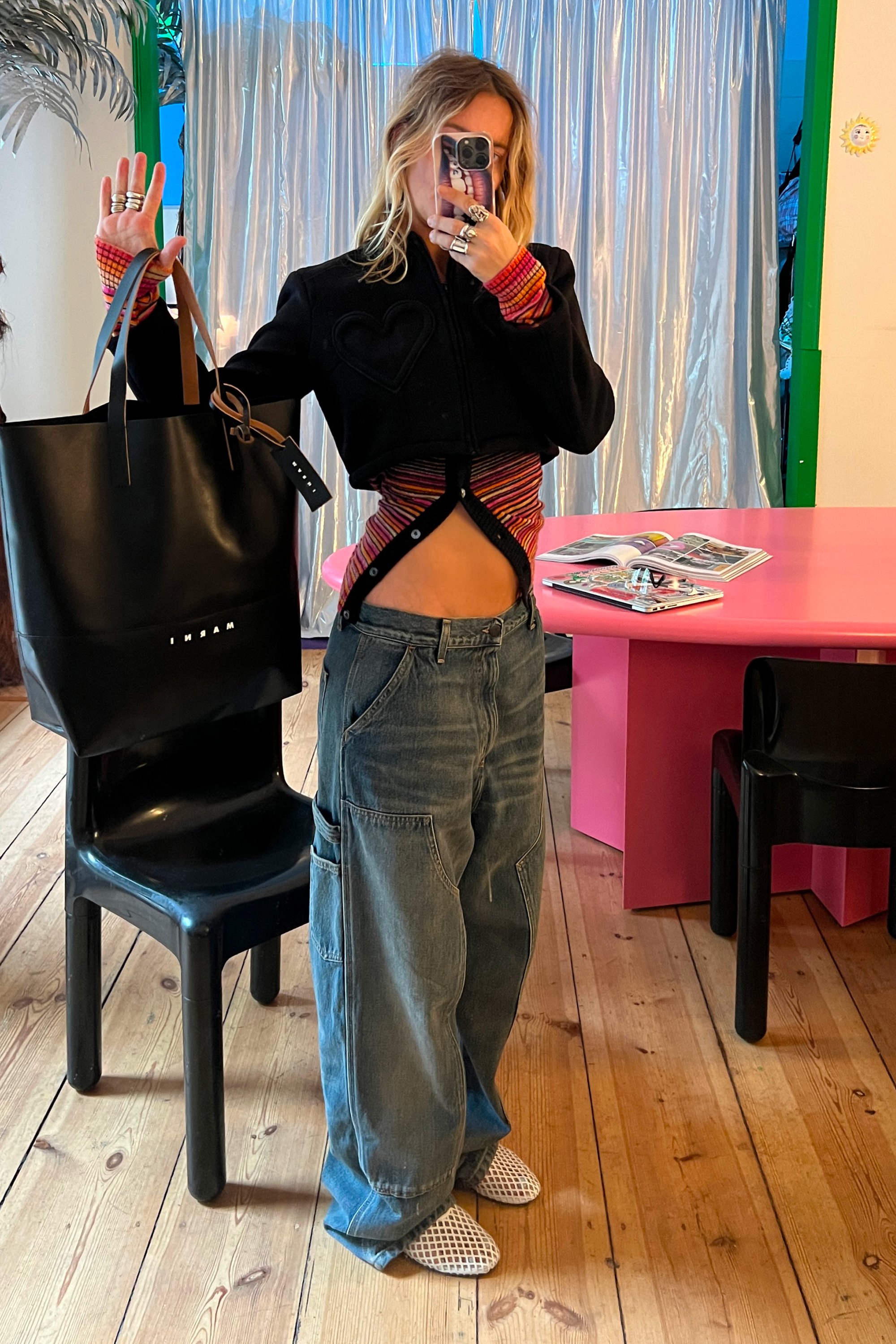 Persona tomando un selfie en el espejo, vistiendo una chaqueta negra corta, una camiseta de rayas coloridas, jeans holgados y zapatillas blancas de rejilla, sosteniendo un gran bolso negro en una habitación luminosa.