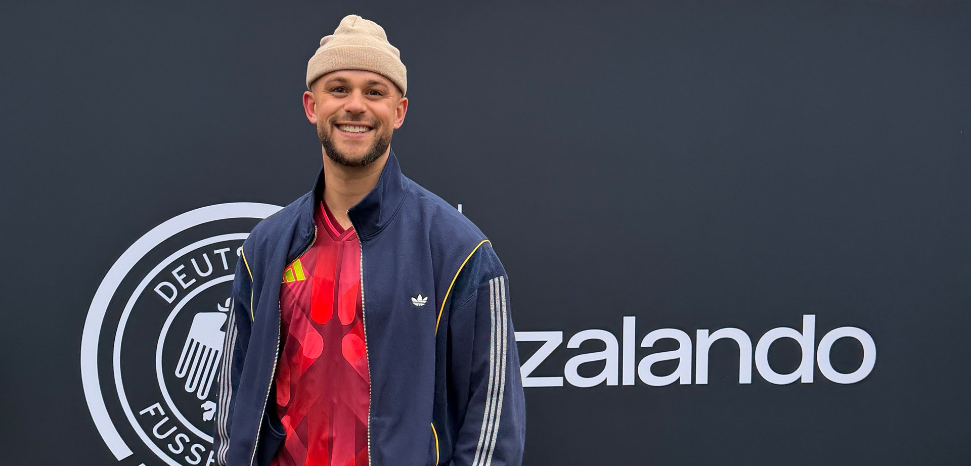 Lächelnder Mann mit beigem Beanie, rotem Sporttrikot und marineblauer Jacke steht vor einem dunklen Hintergrund mit Zalando-Logo.