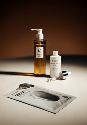 Aceite limpiador en una botella ámbar transparente con un dispensador blanco, botella de sérum blanca con gotero y paquete de papel metálico con detalles del producto.