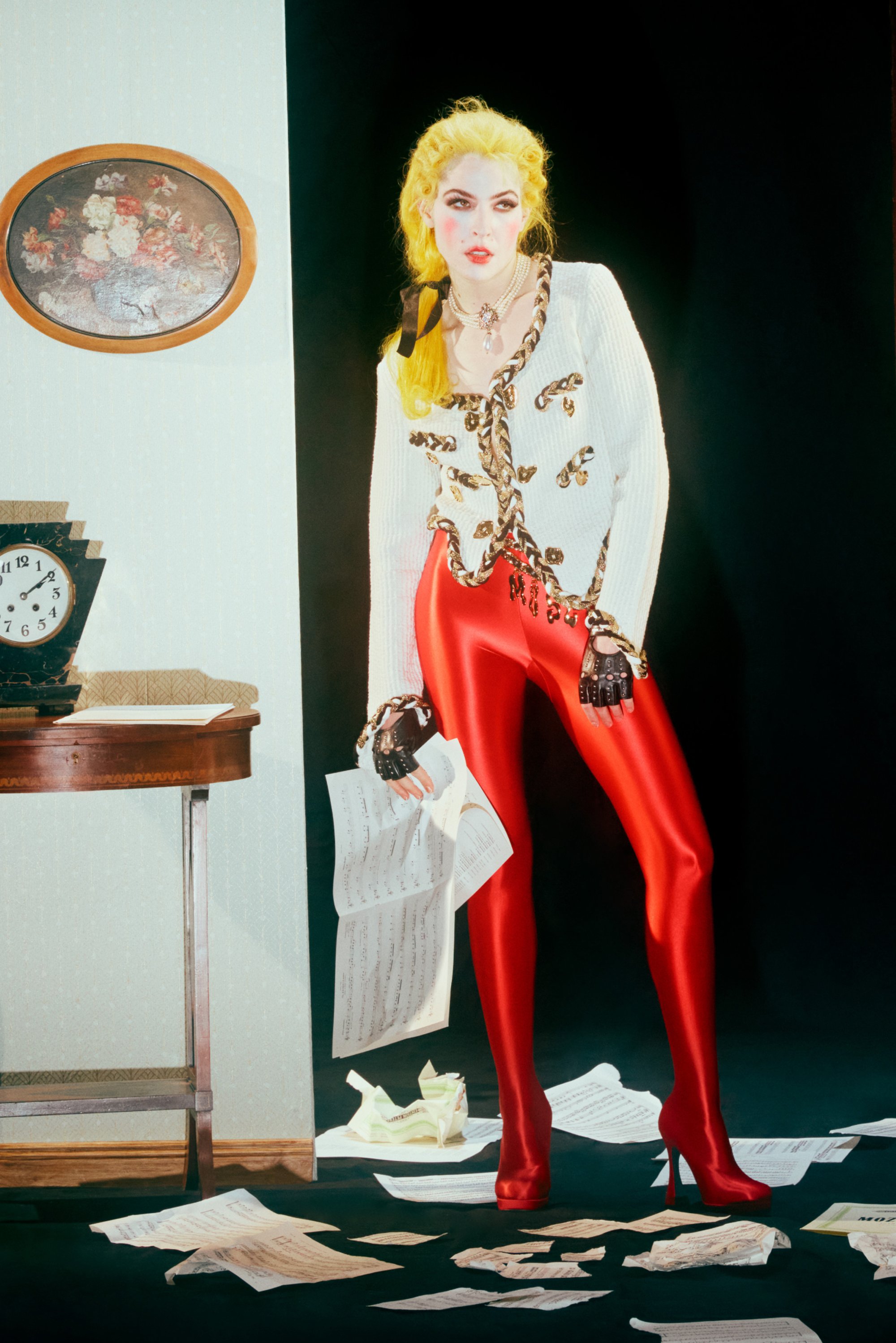 Una persona con cabello amarillo brillante lleva puesto un suéter blanco y leggings rojos, sosteniendo partituras, rodeada de papeles desordenados y un reloj.