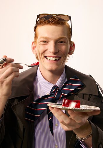 Junge Person mit roten Haaren und Sonnenbrille auf dem Kopf lächelt, während sie einen Teller hält und rote Gelatine mit einer Gabel isst.