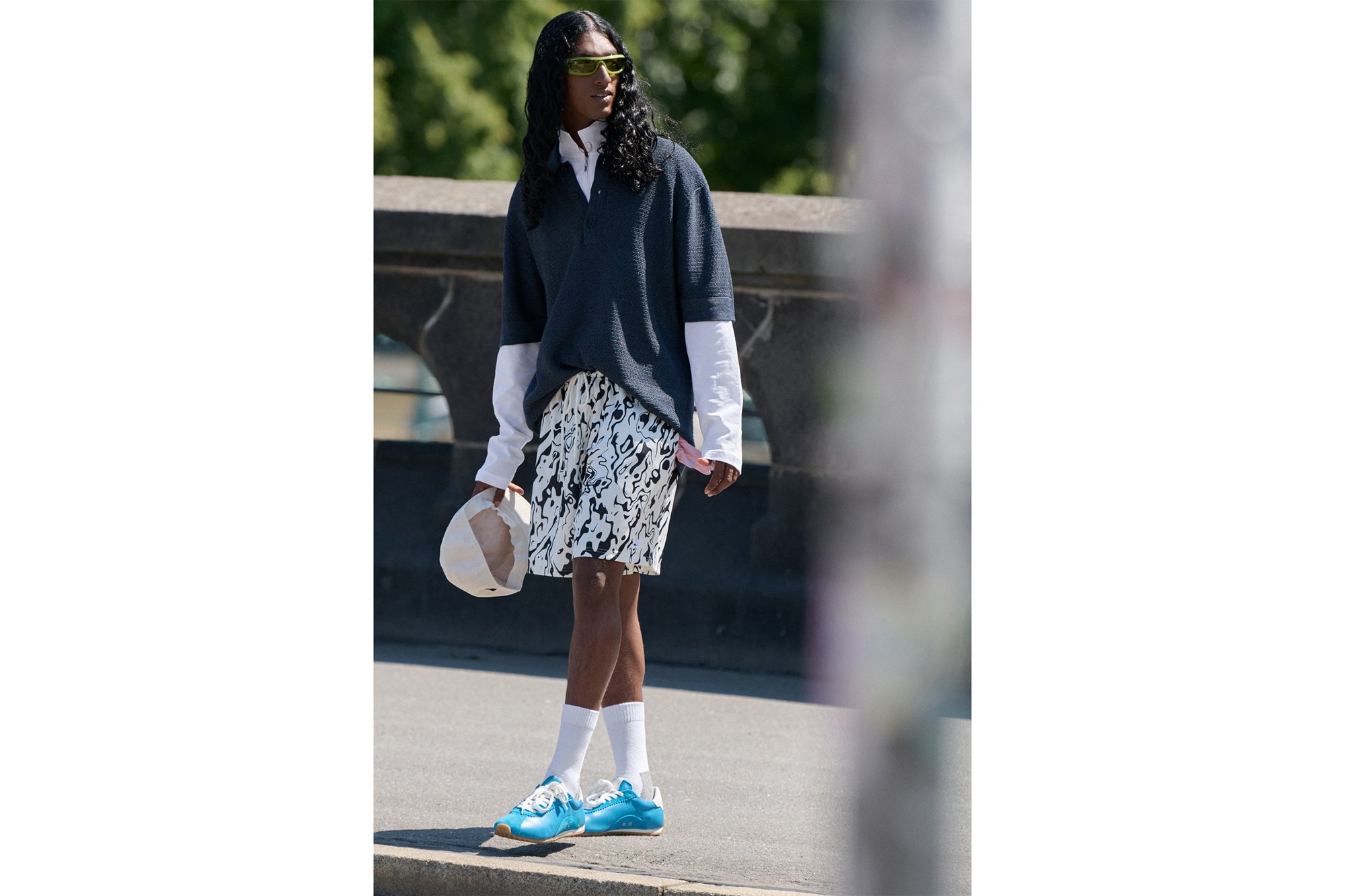 Una persona está de pie en una calle, vistiendo un suéter gris de gran tamaño, una camisa blanca, pantalones cortos con estampado y zapatillas azules, sosteniendo un sombrero.