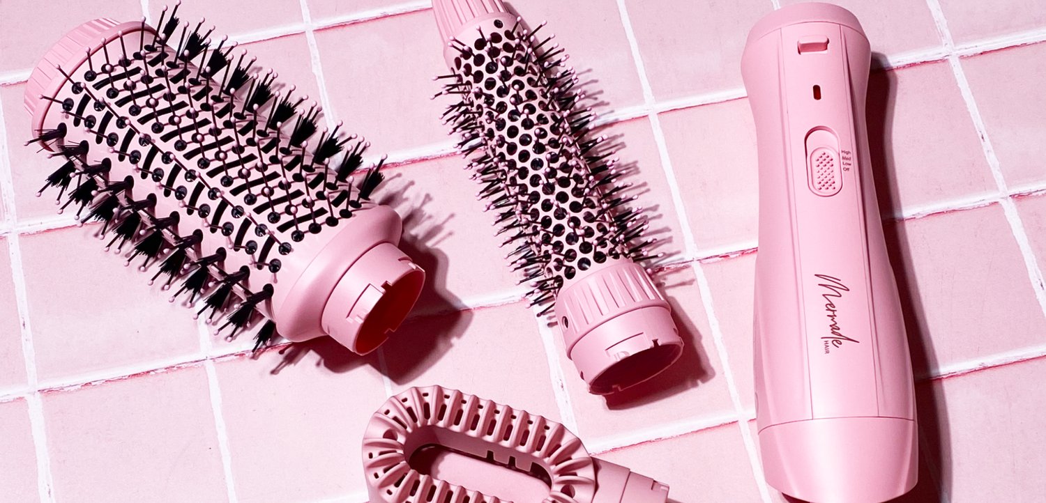 Ensemble d’outils de coiffure roses comprenant deux brosses rondes, une poignée et un accessoire flexible sur une surface carrelée rose.