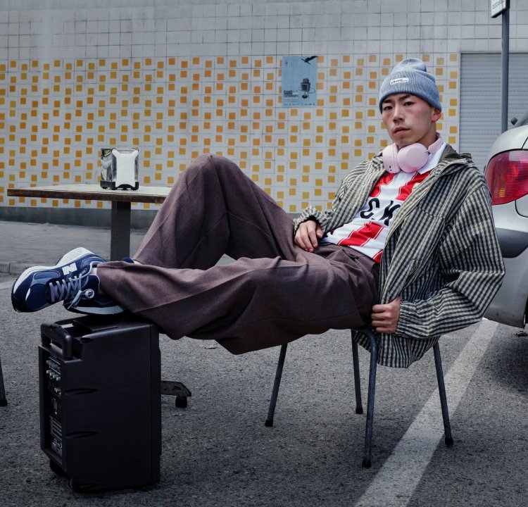 Model wearing a grey striped jacket, a red and white striped shirt, brown trousers, blue trainers, and pink headphones, seated casually on a chair.