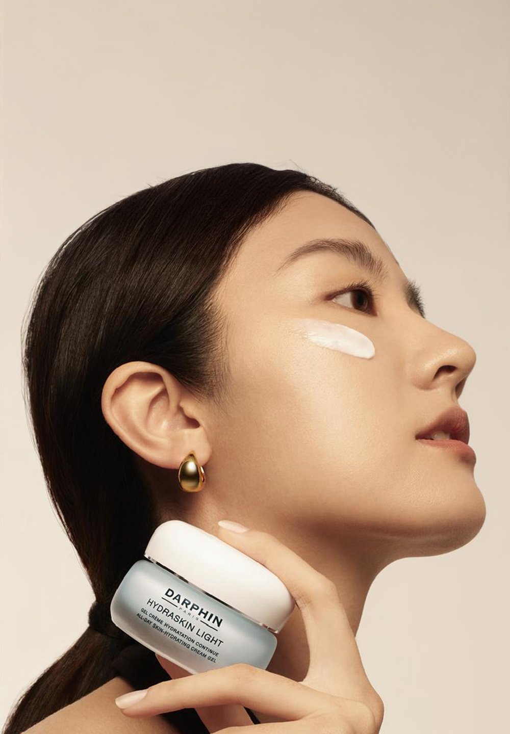 A woman with long dark hair applies moisturizer to her cheek while holding a jar of Darphin Hydraskin Light. She wears gold earrings.