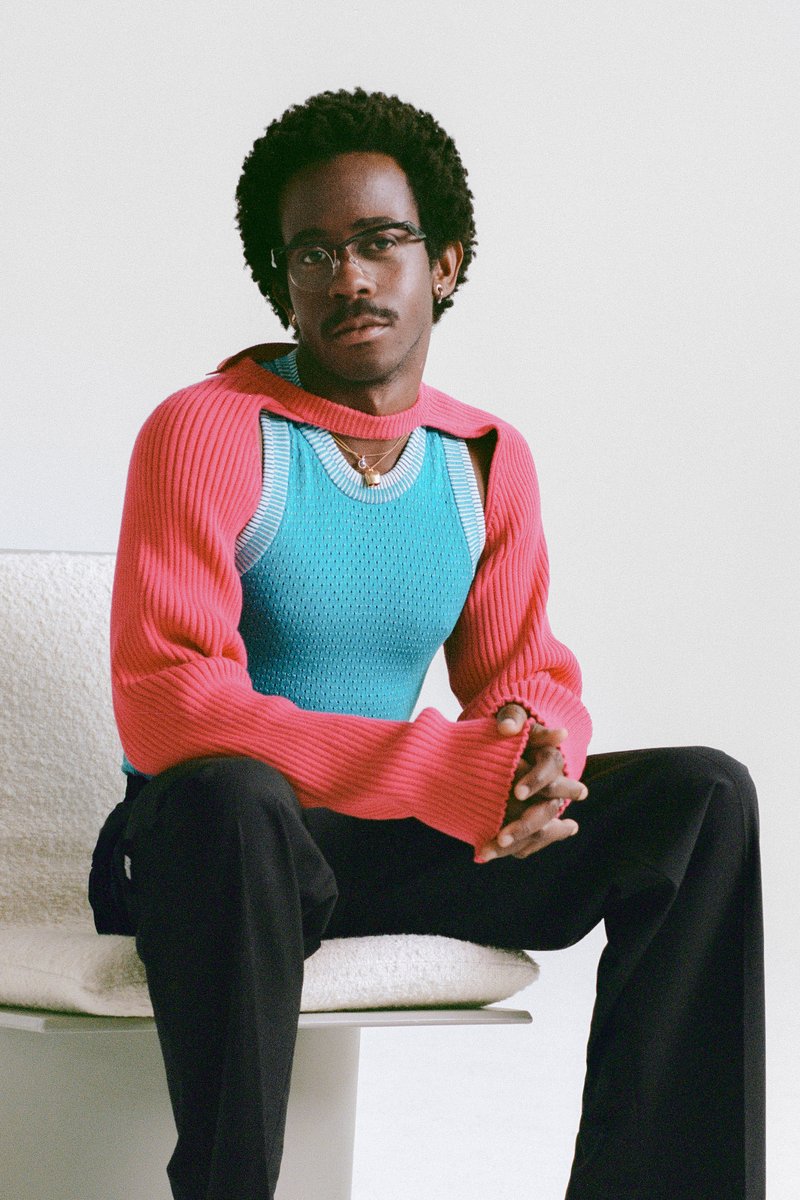 Une personne aux cheveux bouclés est assise sur une chaise de couleur claire, portant un pull coloré avec des manches roses et un corps bleu, les mains jointes.