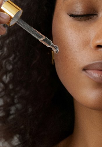 Klar serumpipett med ett gyllene lock som släpper en droppe på slät hud. Texturerat hår är delvis synligt, med en mjuk, naturlig hudton.