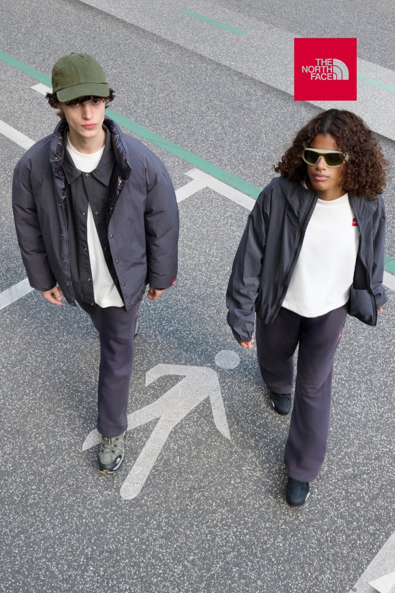 Due persone camminano su una superficie pavimentata. Il primo indossa un giaccone scuro oversize, una camicia bianca e dei pantaloni grigi. Il secondo ha un giubbotto grigio, una maglietta bianca e pantaloni scuri.