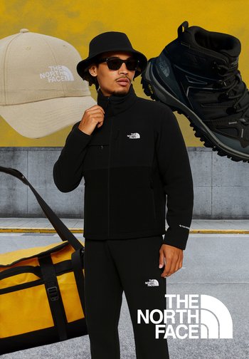 Veste zippée noire et pantalon avec logo, superposés sur un t-shirt noir. Bottes de randonnée noires, sac de sport jaune et casquette kaki en arrière-plan.