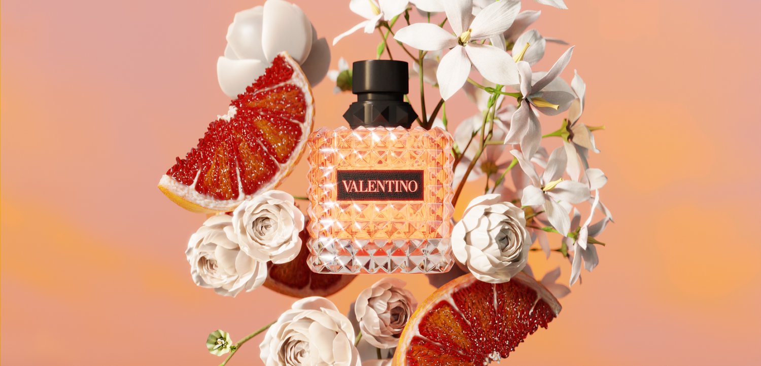 Valentino parfumfles omringd door witte bloemen en gesneden rode grapefruit op een perzikkleurige achtergrond.