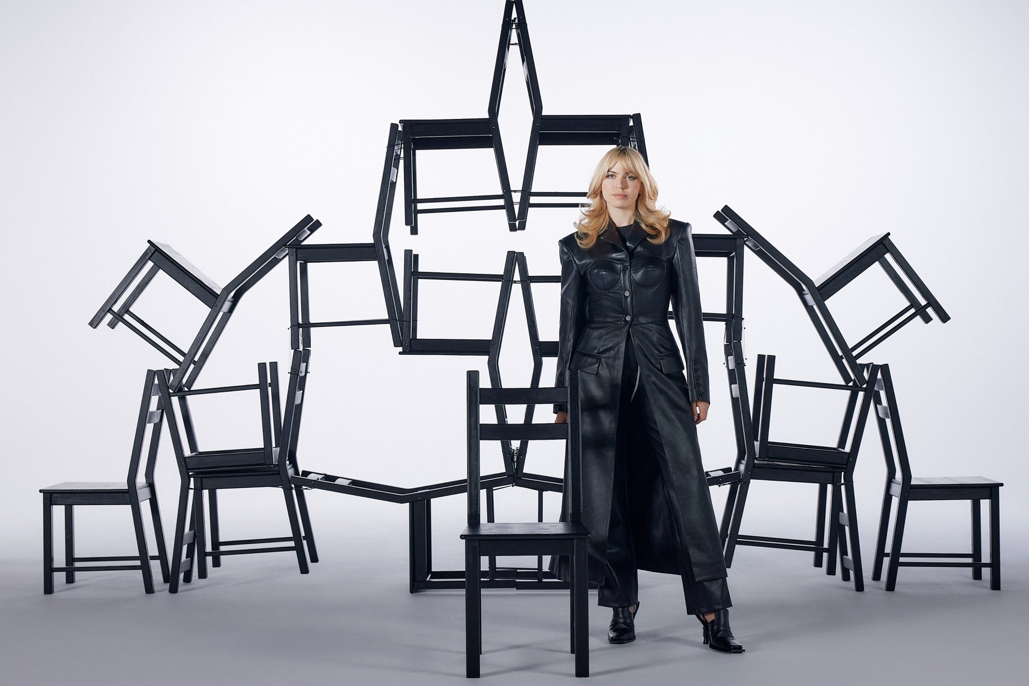 Una silla negra de cuero con un diseño minimalista está rodeada de sillas de madera negras apiladas. La escena presenta una estética moderna y estructurada.