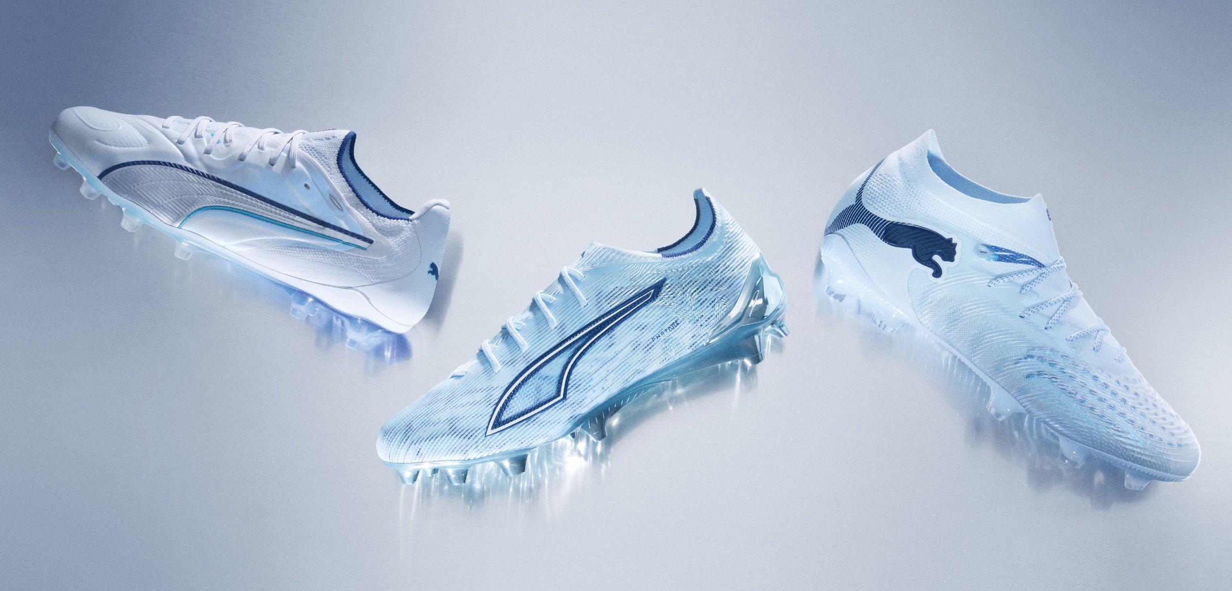 Trois chaussures de football blanches Puma avec des logos bleus et des crampons, exposées sur une surface grise claire réfléchissante.