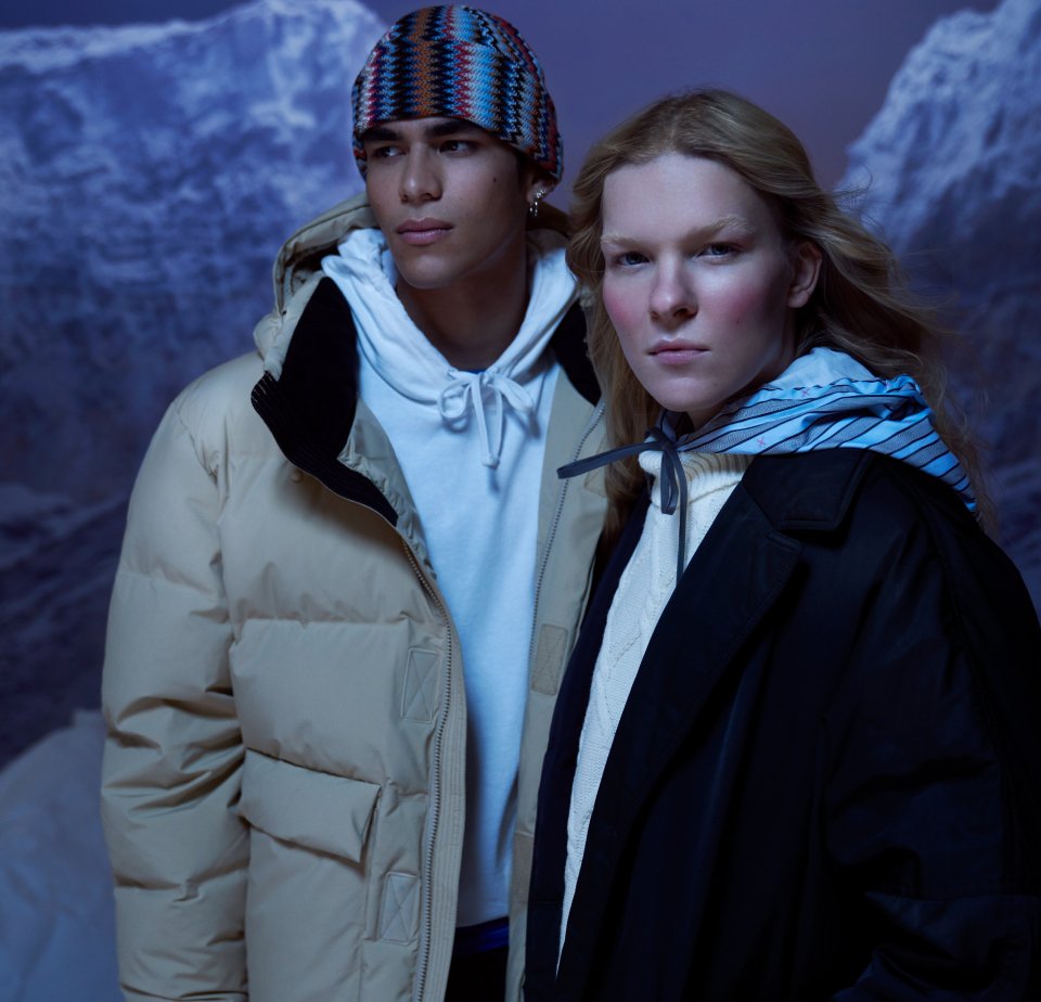 Two models stand in a snowy landscape. One wears a beige puffer jacket and a white hoodie; the other wears a black coat and a blue-striped scarf.