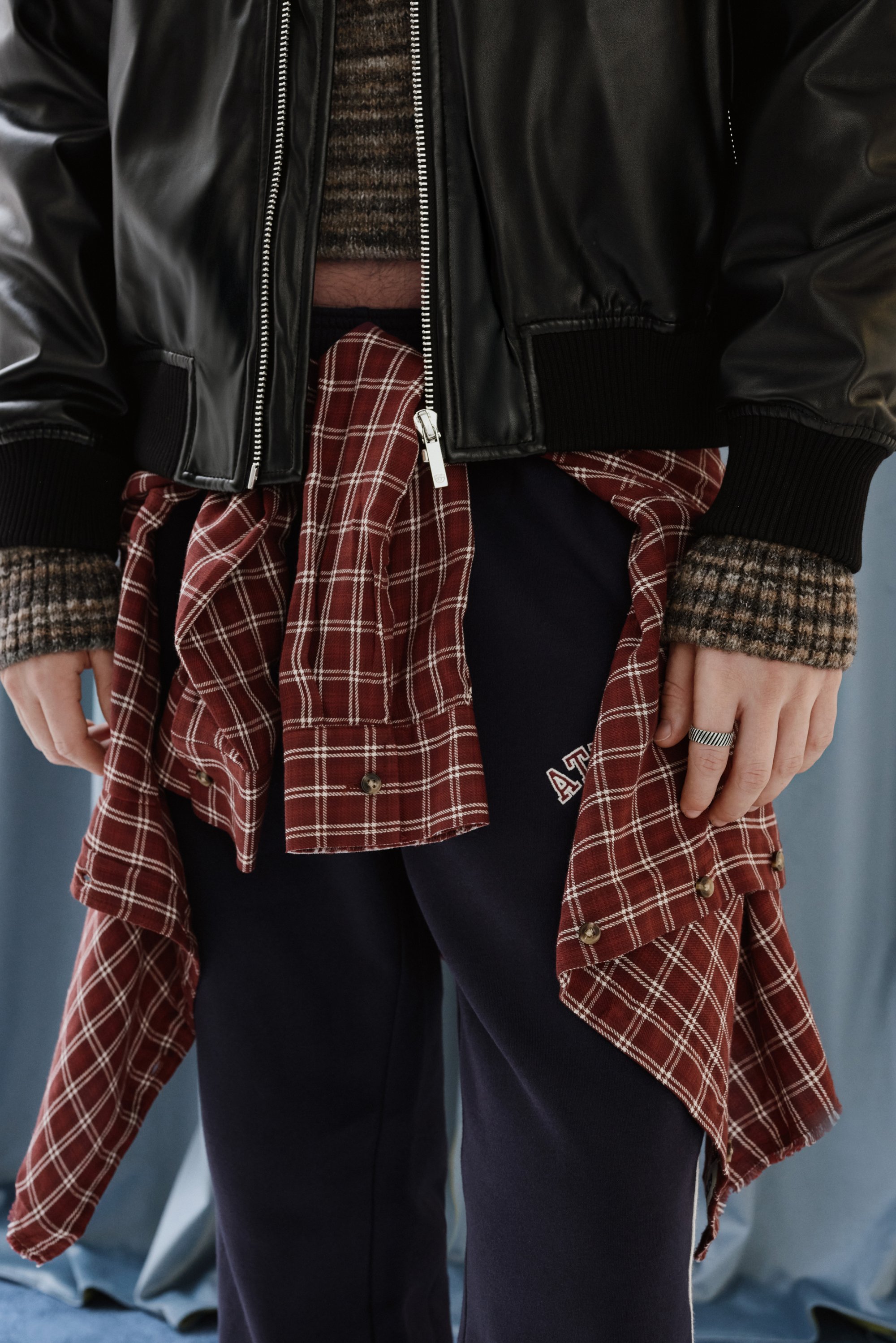 Una persona lleva una chaqueta bomber negra sobre un suéter de rayas marrón, con una camisa de cuadros roja atada a la cintura y pantalones de chándal azul marino.