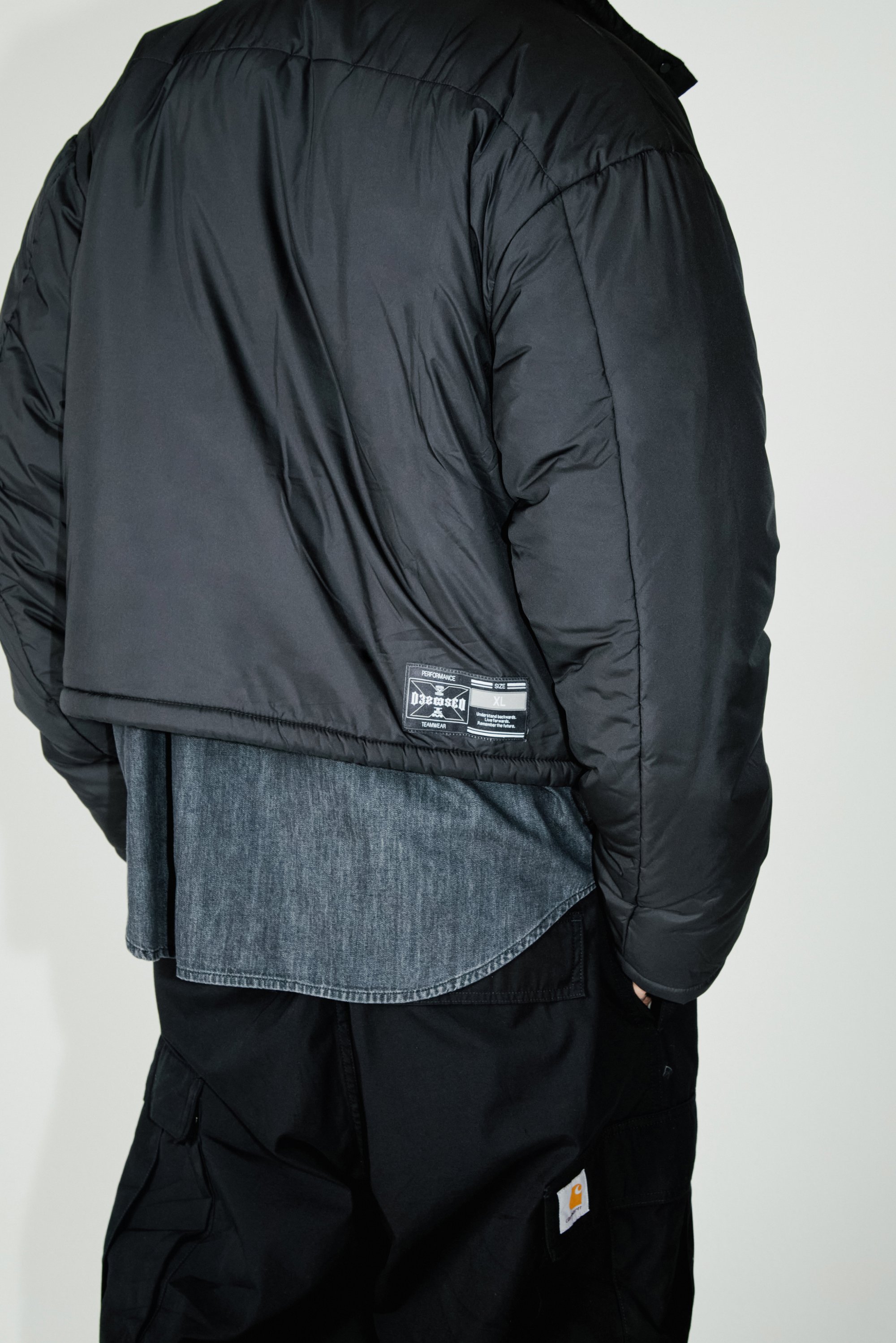 Una persona está de espaldas a la cámara, vistiendo una chaqueta acolchada negra corta sobre una camisa de mezclilla y pantalones cargo negros.