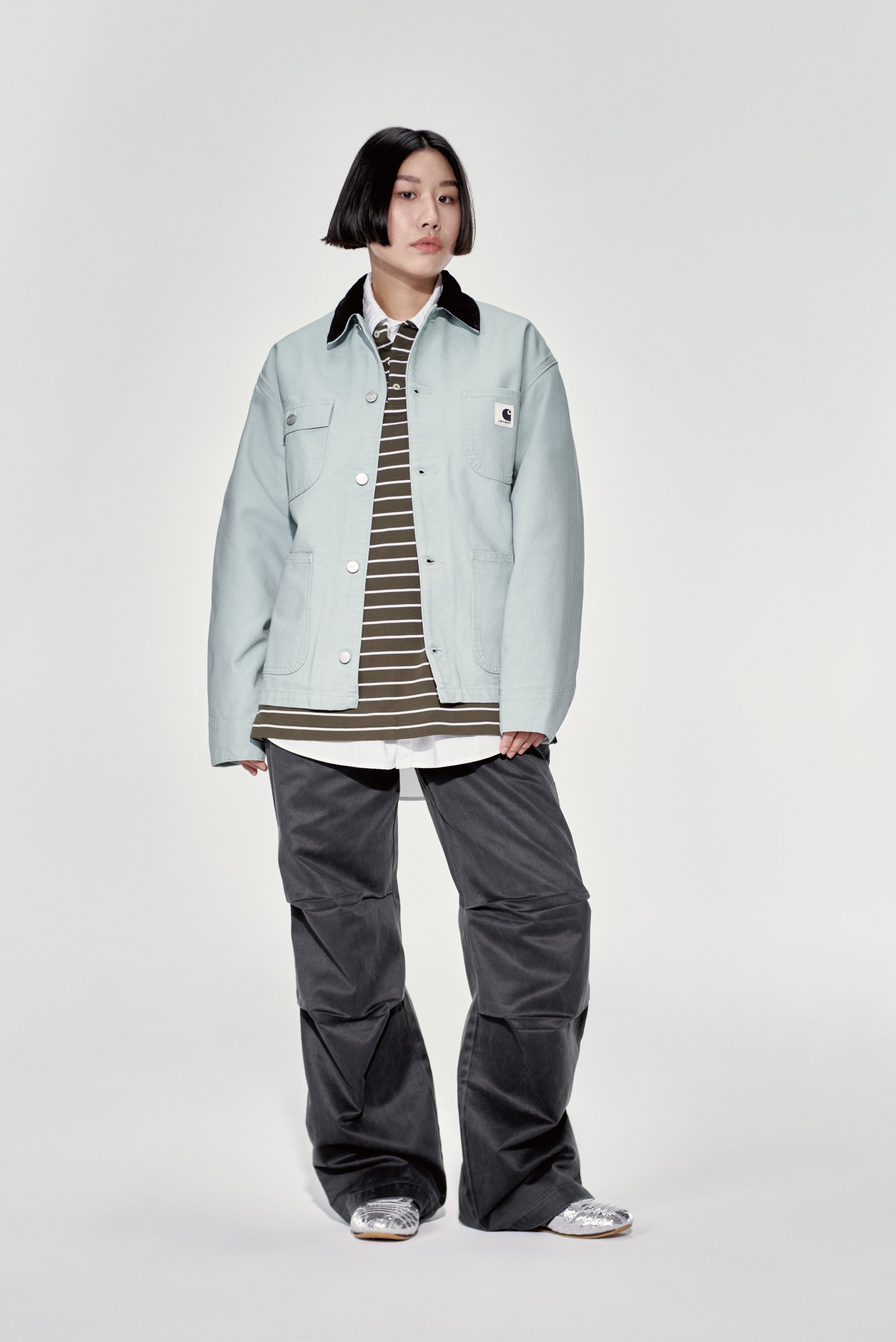 A young person stands confidently, wearing a light blue jacket over a striped shirt, paired with dark baggy pants and silver shoes.