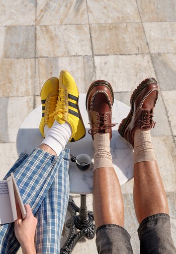 Two pairs of legs resting on a round table, one wearing yellow trainers and checked trousers reading a book, the other with brown boots and rolled-up trousers.
