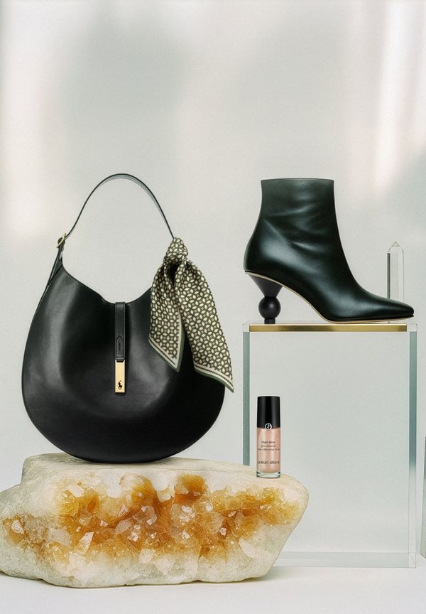 Black leather handbag with a patterned scarf, green ankle boot with a distinctive heel, and a foundation bottle placed on a crystalline stone base.