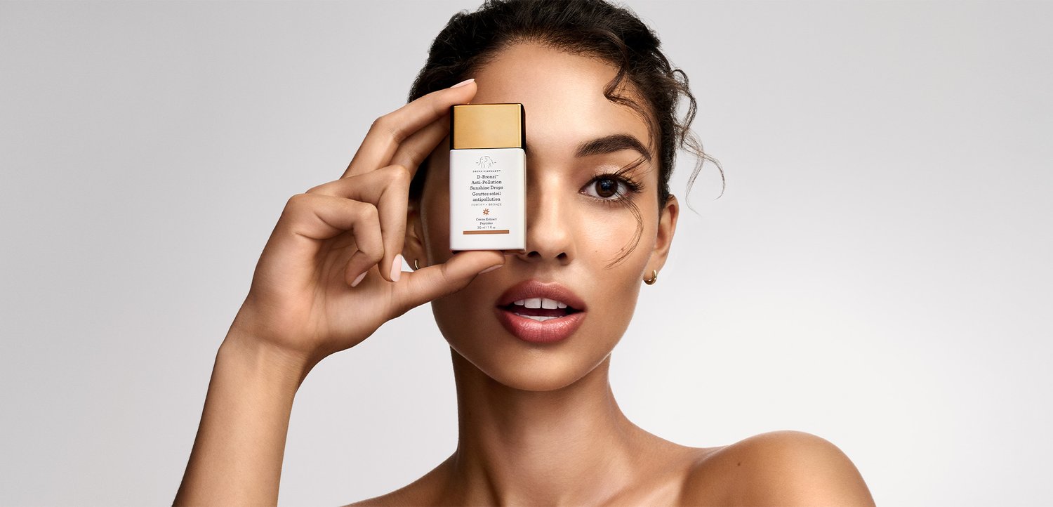 Young woman with clear skin holds a small skincare product bottle near her eye against a plain light background.
