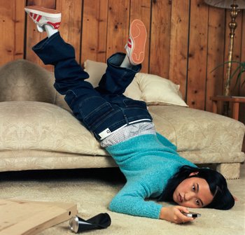 Eine Person liegt auf dem Boden neben einem Sofa, die Beine auf dem Möbelstück und hält eine Fernbedienung in der Hand, gekleidet in lässiger Kleidung und mit einem konzentrierten Gesichtsausdruck.