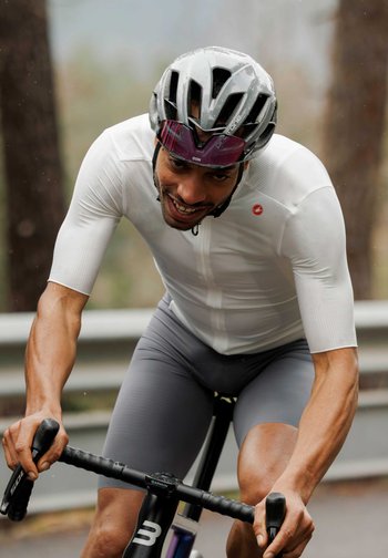 An athletic cyclist grins while pedaling in the rain, wearing a white shirt and gray shorts, with a sleek helmet and glasses. Trees in the background.