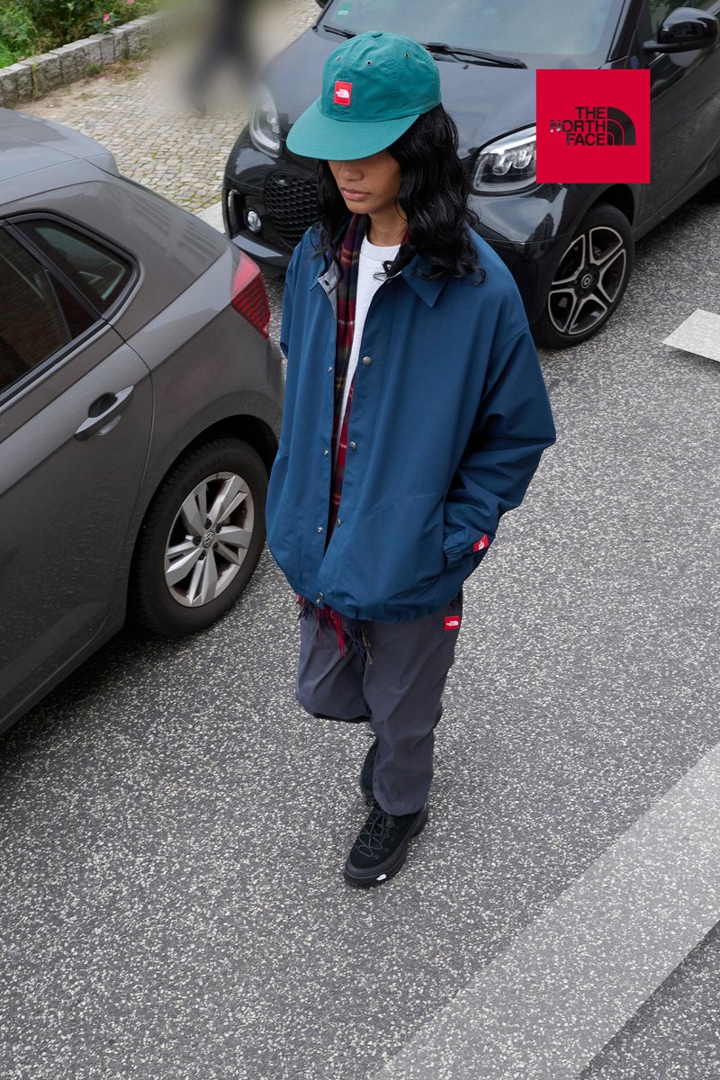 Persoon met een turquoise pet, een blauwe jas en grijze broek loopt tussen geparkeerde auto's op een straat met een rood The North Face-logo.