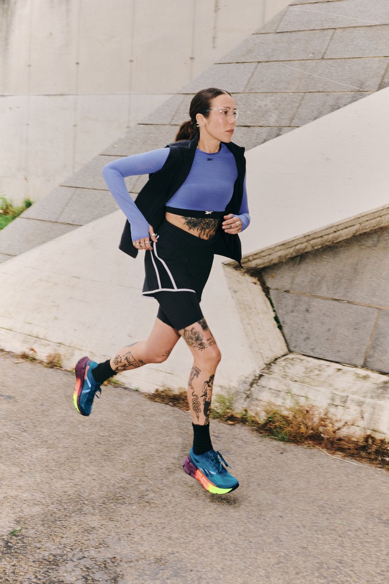 Una mujer con tatuajes corre por un sendero, vistiendo una camiseta de manga larga azul, pantalones cortos negros y zapatillas deportivas coloridas, con un fondo geométrico.