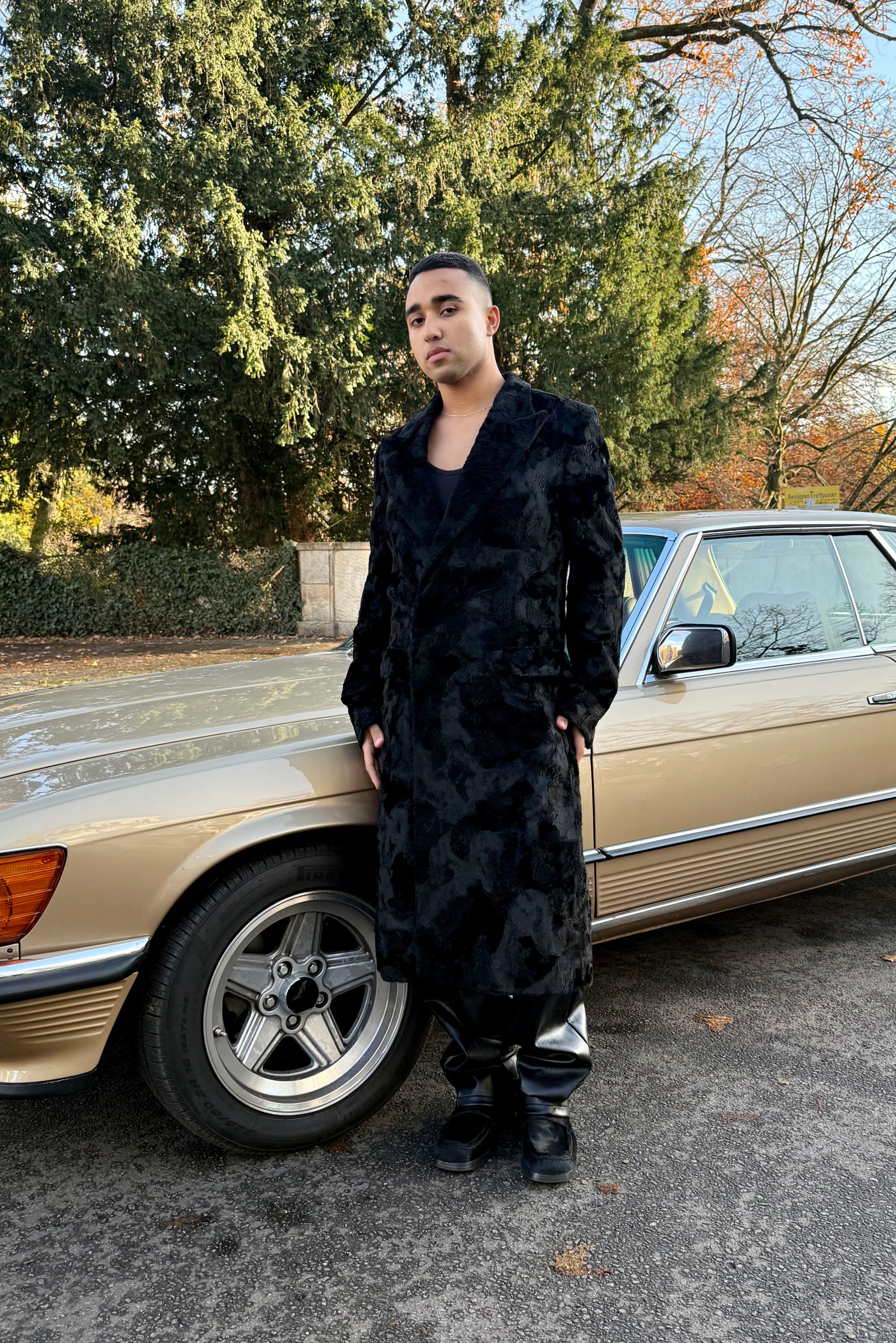 Una persona standing accanto a un'auto d'epoca, indossando un lungo cappotto nero e pantaloni neri, sullo sfondo di alberi e fogliame autunnale.
