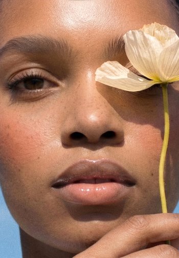 Close-up of a person holding a pale yellow flower near their right eye, highlighting smooth skin, natural makeup, and relaxed facial expression.