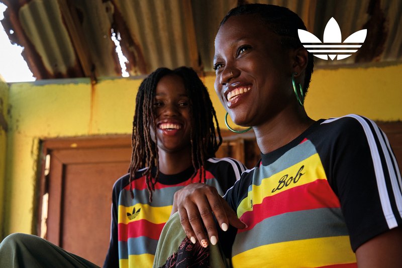 Two smiling women wearing colorful Adidas shirts sit indoors under a corrugated metal roof.