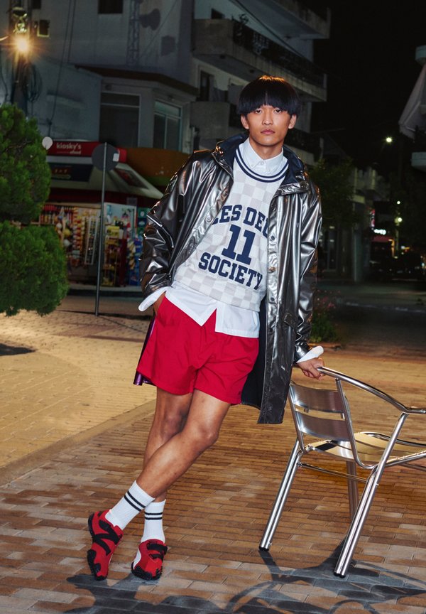 Νεαρός με λαμπερό μαύρο αδιάβροχο, λευκό πουλόβερ, κόκκινα σορτς και κόκκινα παπούτσια, στηρίζεται σε μια μεταλλική καρέκλα σε πεζοδρόμιο πόλης τη νύχτα.