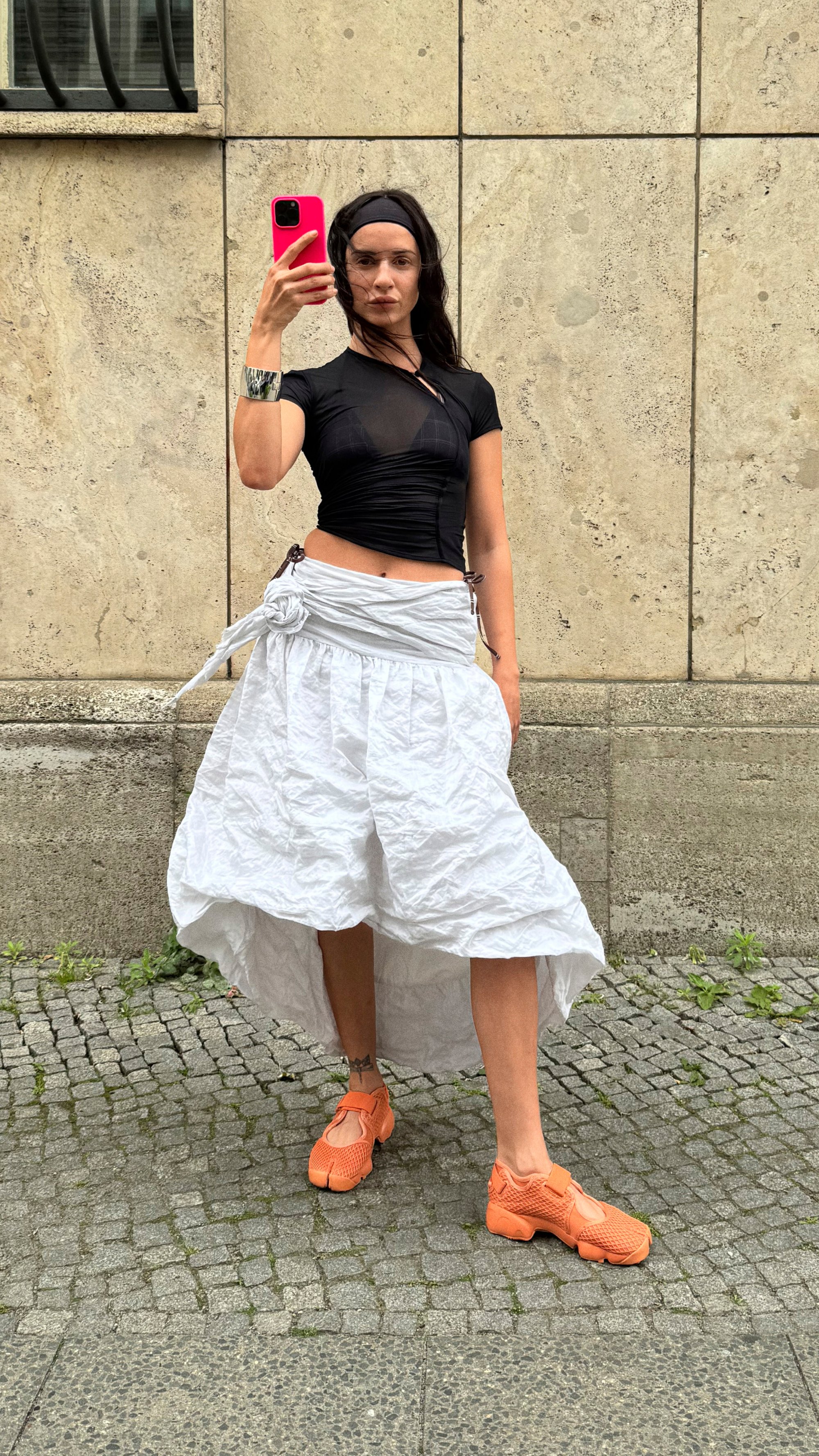 Una persona está al aire libre, vistiendo una blusa negra y una falda blanca, sosteniendo un teléfono rosa mientras se toma una selfie. Se puede ver el suelo de adoquines.