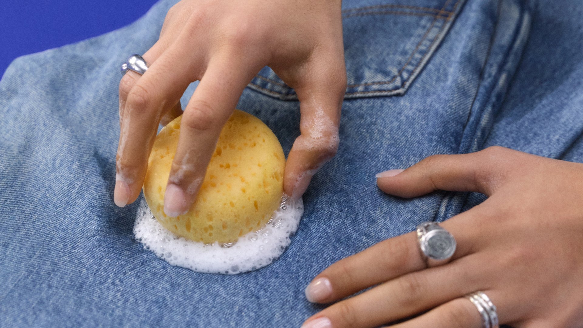 A hand presses a yellow sponge onto a pair of denim jeans, producing bubbles, with rings visible on the fingers.