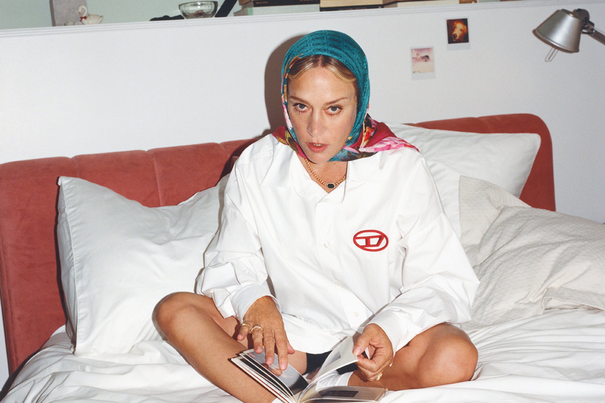 Una donna è seduta a gambe incrociate su un letto, indossa una camicia bianca e una sciarpa colorata, concentrata su un libro fotografico che tiene tra le mani.